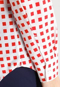Valkoinen kangas, jossa on punainen neliökuvio pitkähihaisessa paidan hihassa ja hihassa tummansinisten housujen päällä.