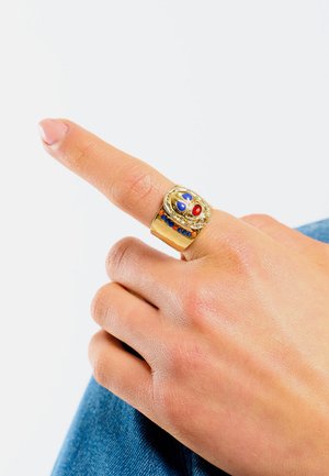 Bague en métal doré avec un large bandeau, présentant un motif en relief avec des pierres rouges et bleues. Texture lisse, accents détaillés et finition polie.