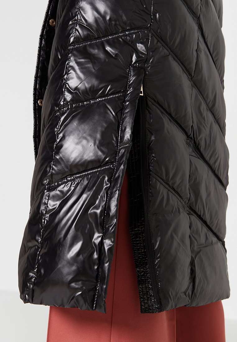 Giubbotto a vento nero con finitura lucida, caratterizzato da sezioni trapuntate prominenti e una zip laterale. Il tessuto è liscio e riflettente.