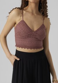 Kvinna klädd i en mauve-färgad spetsig kort topp med tunna band och högmidjade, lösa svarta byxor, hand i fickan, armbandsknapp på handleden.