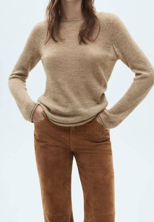 Kvinde iført en beige langærmet sweater og brune ruskindsbukser med hænderne i lommerne mod en ensfarvet lys baggrund.