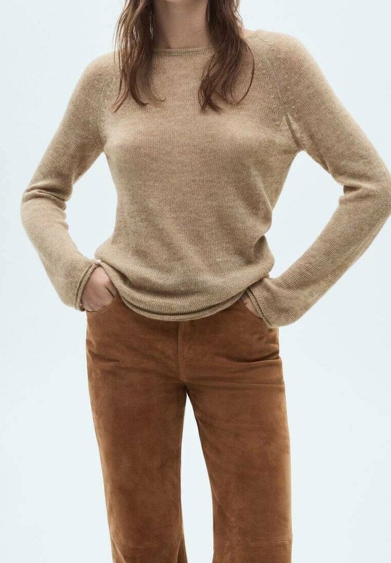 Femme portant un pull à manches longues beige et un pantalon en daim marron, les mains dans les poches, sur un fond clair uni.