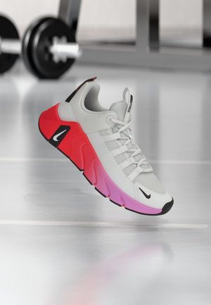 White and gray sneaker with pink and red sole, black Nike logos, floating above a shiny gym floor with blurred weights in the background.