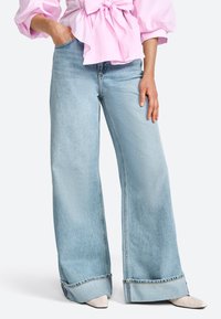 Jeans a gamba larga di un blu chiaro, realizzati in denim, con orlo grezzo, texture leggermente sbiadita, abbinati a una maglietta rosa chiaro e scarpe beige con punta.