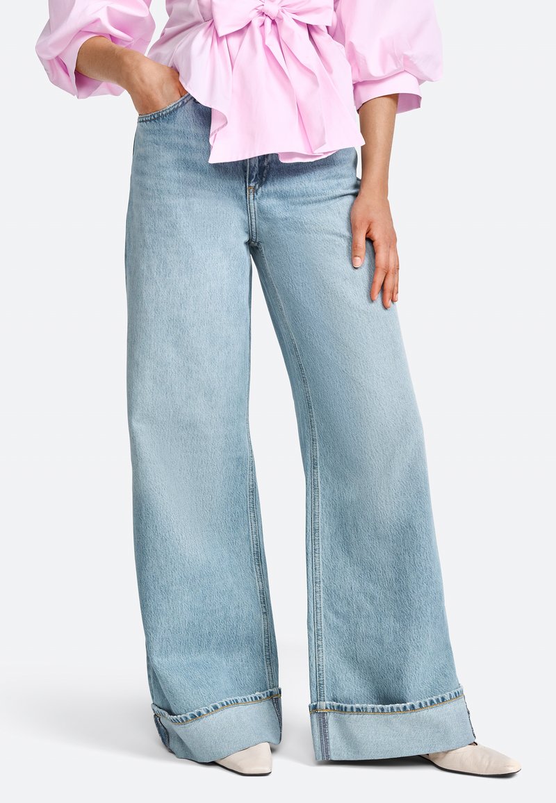 Jeans a gamba larga di un blu chiaro, realizzati in denim, con orlo grezzo, texture leggermente sbiadita, abbinati a una maglietta rosa chiaro e scarpe beige con punta.