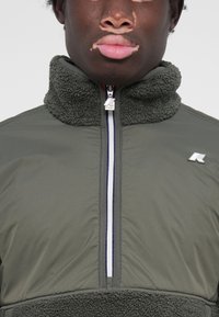 Green fleece jacket with a high collar, zip closure, and silver hardware. Features contrasting white and red stripes along the zipper.