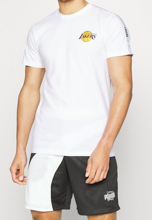 Uomo che indossa una maglietta bianca dei Los Angeles Lakers e pantaloncini Puma neri e bianchi, in piedi con le braccia rilassate.