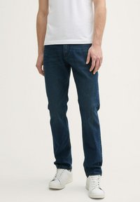 Jeans de denim azul oscuro con diseño de pierna recta, que cuentan con un estilo clásico de cinco bolsillos. Llevados con una camiseta blanca de manga corta y zapatillas blancas.
