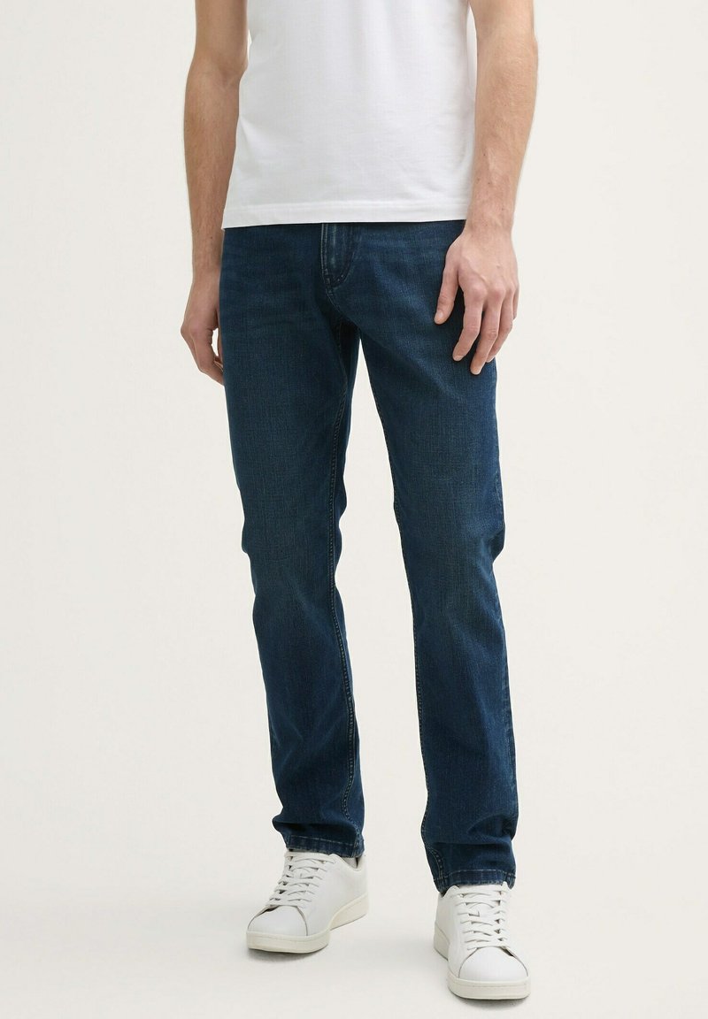 Jeans de denim azul oscuro con diseño de pierna recta, que cuentan con un estilo clásico de cinco bolsillos. Llevados con una camiseta blanca de manga corta y zapatillas blancas.