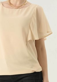 Beige chiffon top met korte, vloeiende mouwen. Heeft een ronde hals en een zachte textuur. Gecombineerd met slanke zwarte broeken.