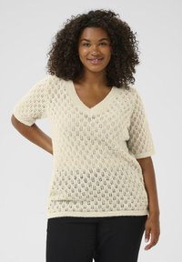 Glimlachende vrouw met krullend haar in een crèmekleurige gebreide trui met korte mouwen en een bladdessin, in zwarte broek, staand tegen een effen achtergrond.