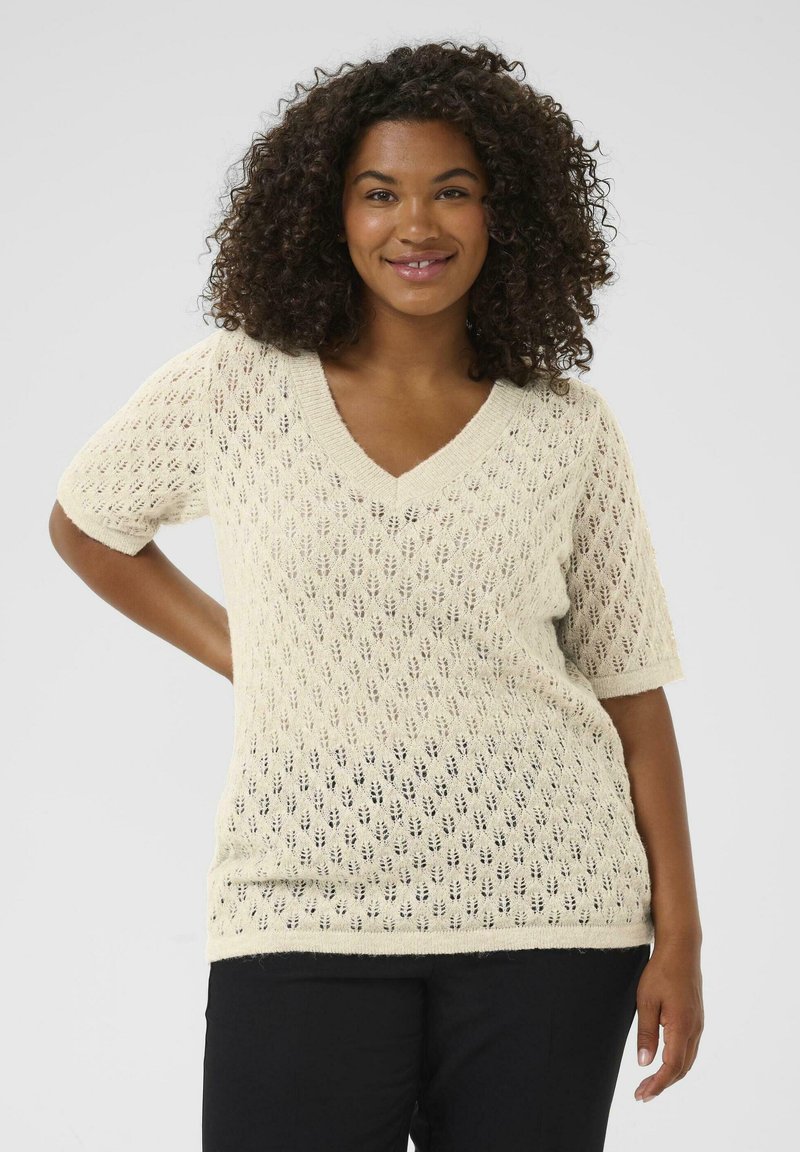Glimlachende vrouw met krullend haar in een crèmekleurige gebreide trui met korte mouwen en een bladdessin, in zwarte broek, staand tegen een effen achtergrond.