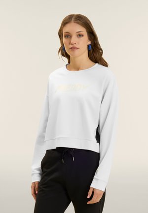 Giovane donna con capelli castani indossa una felpa bianca corta a maniche lunghe e pantaloni neri con coulisse, in piedi su sfondo neutro.