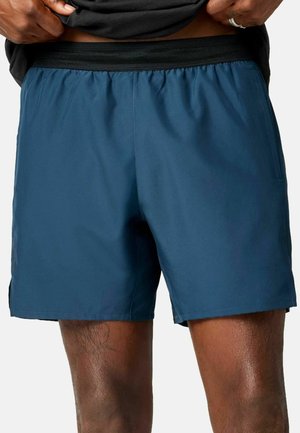 Person trägt dunkelblaue Sportshorts mit elastischem Bund und hält ein schwarzes Shirt hoch, um die Shorts zu zeigen.