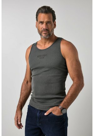 Mann mit Bart, der ein graues ärmelloses Tanktop und dunkelblaue Jeans trägt, steht mit einer Hand in der Tasche vor einem einfarbigen Hintergrund.
