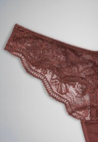 Tissu en dentelle marron avec motifs floraux et bordures festonnées, partie d'un vêtement de lingerie sur un fond gris clair uni.
