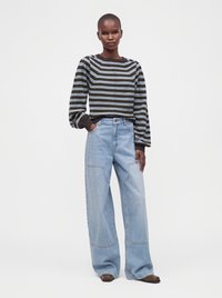 Femme portant un pull rayé à manches longues et un jean bleu clair à jambes larges, debout avec une main dans la poche devant un fond uni.