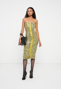 Arany flitteres ruha, pánt nélküli fazonnal, középen sárga csíkkal és testhez álló siluettel. A modell fekete kézitáskát tart és fekete magassarkú cipőt visel.