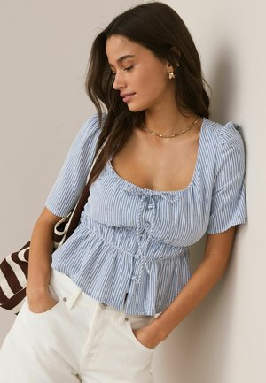 Kobieta z długimi ciemnymi włosami, ubrana w niebiesko-białą pasiastą bluzkę i białe spodnie, stoi z rękami w kieszeniach na tle jasnej ściany.