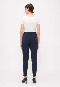 Marineblauwe tapered broek met elastische tailleband, twee achterzakken en subtiele verticale textuur. Gekleed met een strakke witte top en hakken.