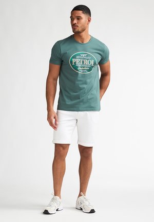 Groen T-shirt met een vintage "PETROL"-opdruk, gecombineerd met witte opgerolde shorts en grijze sportschoenen. Zachte katoenen textuur.