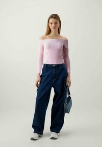Pink off-the-shoulder long-sleeve top paired with loose blue denim jeans. Model holds a small blue bag. White sneakers complete the outfit.