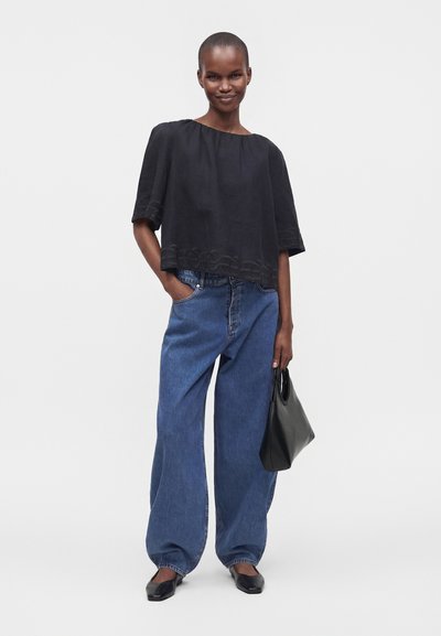 Femme aux cheveux courts portant un haut noir ample, un jean large bleu, des ballerines noires, tenant un sac à main noir, debout devant un fond clair uni.
