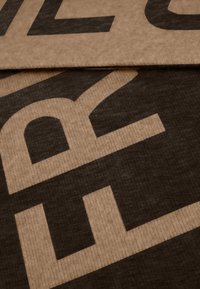 Beige stof met grote zwarte letters. De textuur lijkt glad met een lichte glans, wat een moderne, minimalistische uitstraling benadrukt.