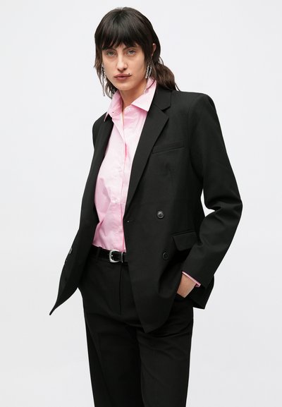 Μαύρο σακάκι διπλόπέτο, ροζ πουκάμισο με κουμπιά, εφαρμοστή γραμμή, υφασμάτινη υφή, με ζώνη και μακριά κρεμαστά σκουλαρίκια, το μοντέλο ποζάρει με αυτοπεποίθηση.