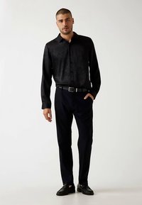 Schwarzes langärmeliges Hemd mit strukturierter Oberfläche, kombiniert mit dunkler Hose und schwarzem Gürtel. Als Schuhe schwarze, gewebte Slipper.