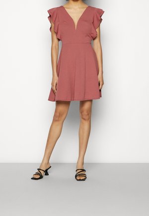 Kobieta ubrana w krótką sukienkę w kolorze pudrowego różu z głębokim dekoltem w kształcie litery V i falbanianymi krótkimi rękawami, w połączeniu z czarnymi sandałami na obcasie z paskami.