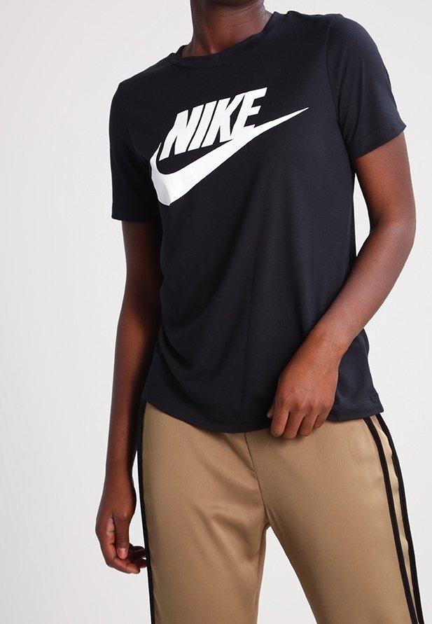 Osoba ubrana w czarną koszulkę Nike z białym logo oraz beżowe spodnie z czarnymi bocznymi paskami, stojąca z jedną ręką dotykającą spodnie.