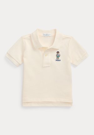 Pikepaita vaaleasta puuvillakankaasta, jossa on taittuva kaulus, lyhyet hihat ja brodeerattu logo rinnassa.