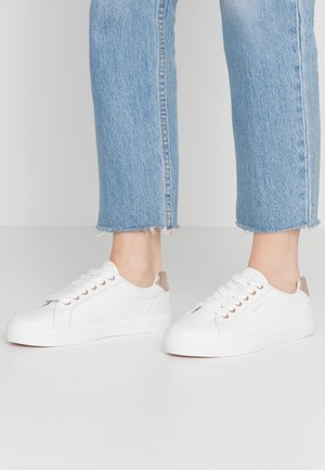 Pieds portant des baskets basses blanches avec des languettes beiges au talon et un jean bleu clair raccourci avec des ourlets effilochés sur un sol blanc.