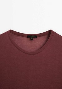 Gros plan d’un t-shirt bordeaux uni à col rond montrant le col et l’étiquette de la marque sur fond blanc.
