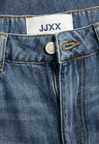 Denim jeans with a zipper fly, featuring a silver button and contrasting orange stitching. A white label displays "JJXX" at the waistband.