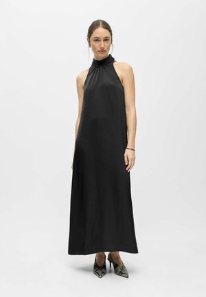 Femme portant une robe maxi noire sans manches à col halter avec des talons hauts à motifs noir et blanc, debout devant un fond blanc uni.