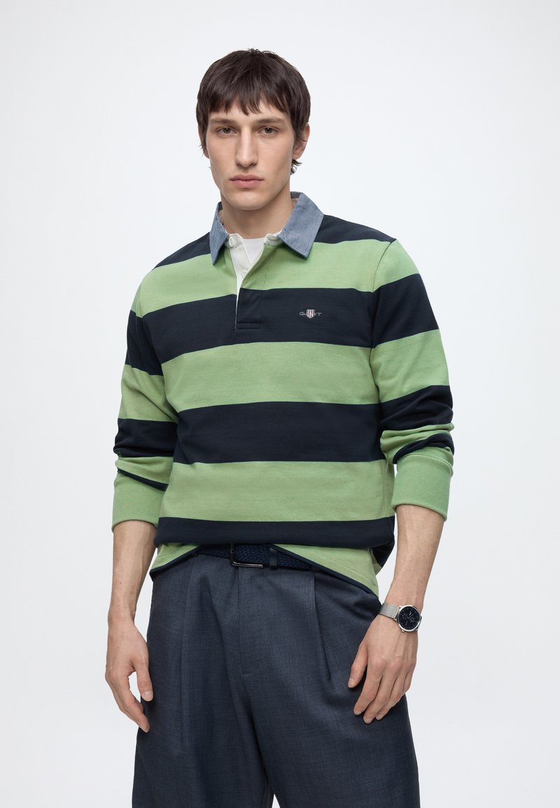Homme debout portant un polo de rugby rayé vert et bleu marine avec un col bleu, un pantalon sombre et une montre, faisant face à l'avant sur un fond uni.