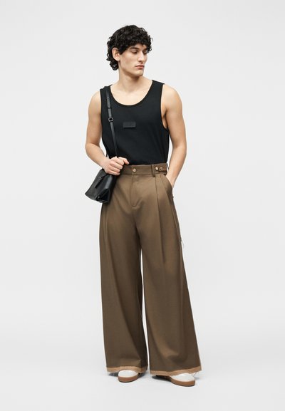 Νεαρός άνδρας που φοράει μαύρο αμάνικο τοπ, φαρδιά καφέ παντελόνια, λευκά παπούτσια και κρατά μια μαύρη τσάντα ώμου, στέκεται μπροστά σε ένα απλό υπόβαθρο.