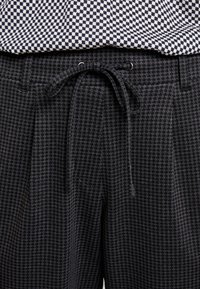 Zwart en grijs geblokte broek met een trekkoord in de taille, voorzien van plooien en een gestructureerde stof. Daaronder zit een patroonblouse in marineblauw en wit.