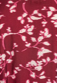 Punainen kangas, jossa on kukkakuosi vaaleanpunaisen ja valkoisen sävyissä, esittää abstrakteja kukkia ja oksia pehmeässä, teksturoidussa pinnassa.