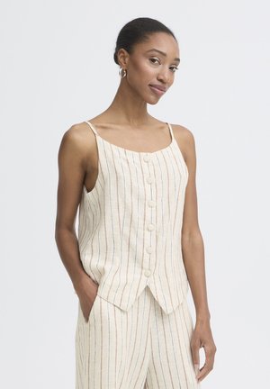 Femeie cu cercei de tip cerc, purtând top bej fără mâneci și pantaloni asortați cu dungi verticale subțiri, stând cu o mână în buzunar.