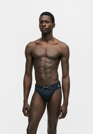 Muskularny mężczyzna stojący, ubrany w ciemnoniebieskie kąpielówki ze sznurkiem z przodu, na jednolitym jasnoszarym tle.