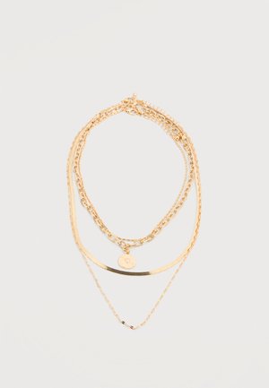 Trzywarstwowy naszyjnik z łańcuszków ze złota o różnych ogniwach, jeden z okrągłą zawieszką z wygrawerowanym wzorem, na jasnym tle.