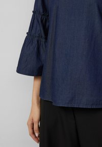 Marineblaue Denim-Bluse mit leicht gerafften Dreiviertelärmel, die einen lockeren Schnitt und eine glatte Textur aufweist, kombiniert mit schwarzen Hosen.