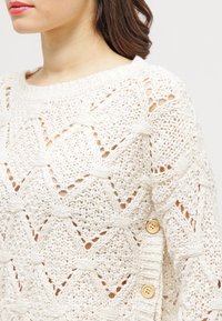 Stickad tröja i krämfärg, med ett diamantmönster, öppna stickdetaljer och träknappsinlägg på sidan.