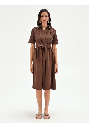 Femme portant une robe marron longueur genou avec un haut en dentelle florale, des manches courtes, une ceinture à nouer à la taille et des sandales plates, se tenant devant un fond uni.