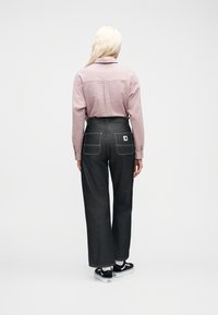 Γυναίκα με μακριά ξανθά μαλλιά, που φοράει ένα ροζ πουκάμισο με υφή, σκούρο τζιν με φαρδιά πόδια και ψηλή μέση, λευκές κάλτσες και ασπρόμαυρα αθλητικά, γυρισμένη με την πλάτη.