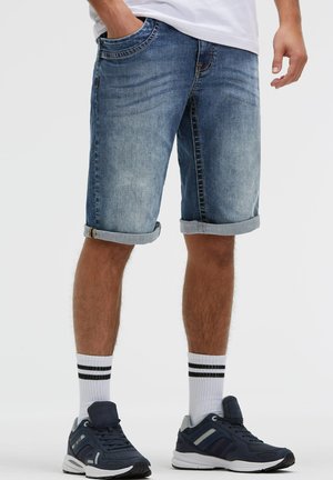 Mann trägt hochgekrempelte blaue Denim-Shorts, weiße Socken mit schwarzen Streifen und dunkelblaue Sneakers, steht mit einer Hand in der Tasche.