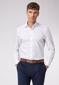 Witte overhemdd met een verspreide kraag, lange mouwen en een knoopsluiting aan de voorkant, gecombineerd met marineblauwe broek en een bruine riem. Soepele textuur.